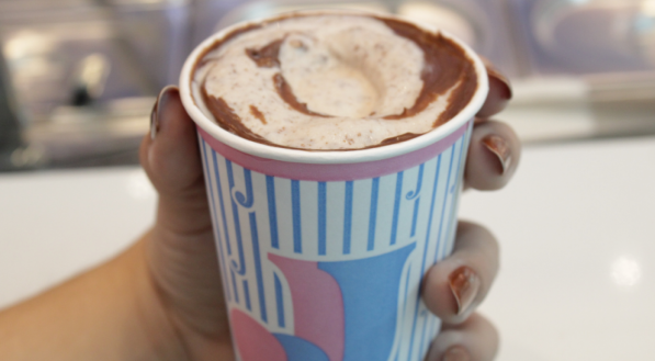 A imagem mostra um milkshake cremoso de Ferrero Rcher no copo listrado da Johnny Joy, sendo segurado por uma mão.