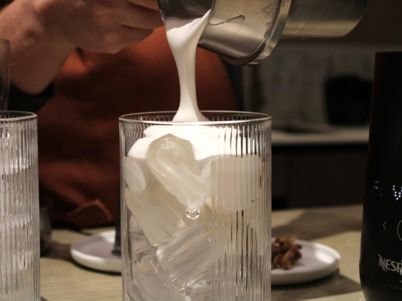 A imagem mostra espuma de leite cremosa sendo despejada de um recipiente de metal em um copo alto cheio de gelo. A imagem mostra espuma de leite cremosa sendo despejada de um recipiente de metal em um copo alto cheio de gelo.