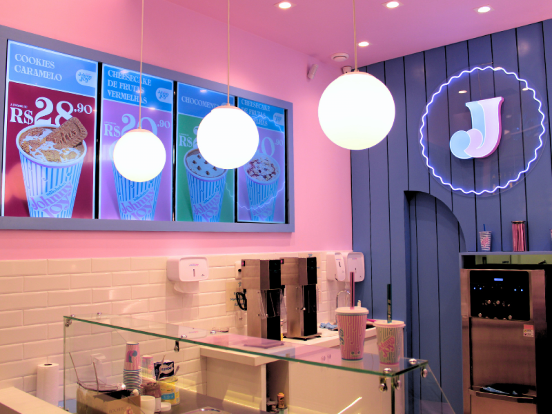 A imagem mostra o interior da loja Johnny Joy em tons de rosa e lilás, com um menu digital de milkshakes atrás do balcão.