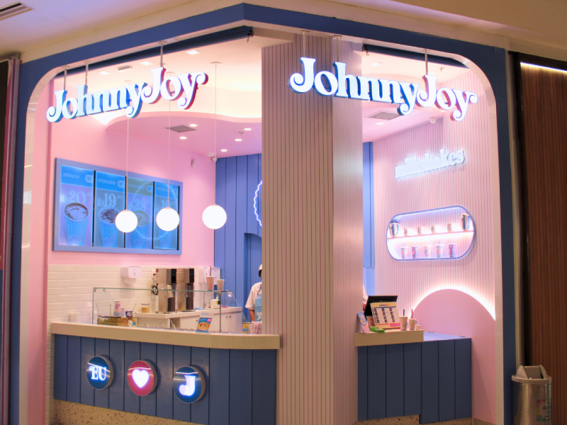 A imagem mostra a fachada da loja Johnny Joy com a logomarca iluminada, cores pastel e ingredientes e copos no balcão.
