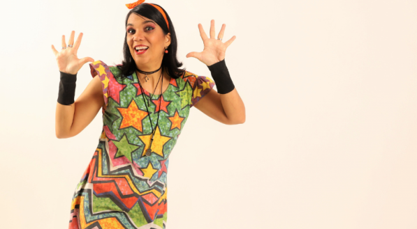 Mulher sorridente com vestido colorido de estampa geométrica e estrelas, levantando as duas mãos abertas. Ela usa uma tiara laranja e munhequeiras pretas, em fundo bege liso.