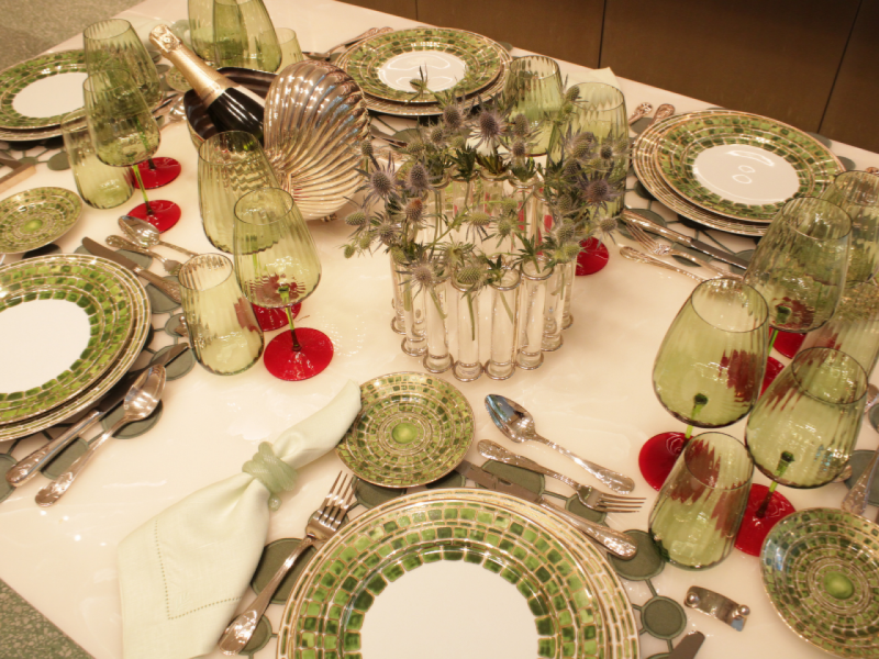 A imagem mostra uma mesa posta com a Coleção Jade, incluindo pratos e taças onduladas, e um arranjo central de flores em um vaso de tubos de ensaio. A imagem mostra uma mesa posta com a Coleção Jade, incluindo pratos e taças onduladas, e um arranjo central de flores em um vaso de tubos de ensaio.