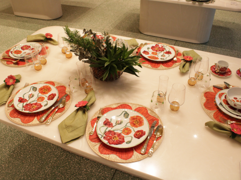 A imagem mostra uma vista de cima da mesa posta para seis, destacando os pratos florais Papoula sobre sousplats laranja e o arranjo de centro em folhagens. A imagem mostra uma vista de cima da mesa posta para seis, destacando os pratos florais Papoula sobre sousplats laranja e o arranjo de centro em folhagens.