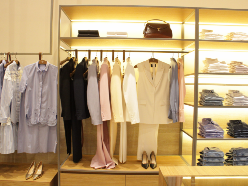 Interior da loja com prateleiras e araras exibindo camisas femininas, ternos claros, e acessórios como sapatos de salto e bolsas. 