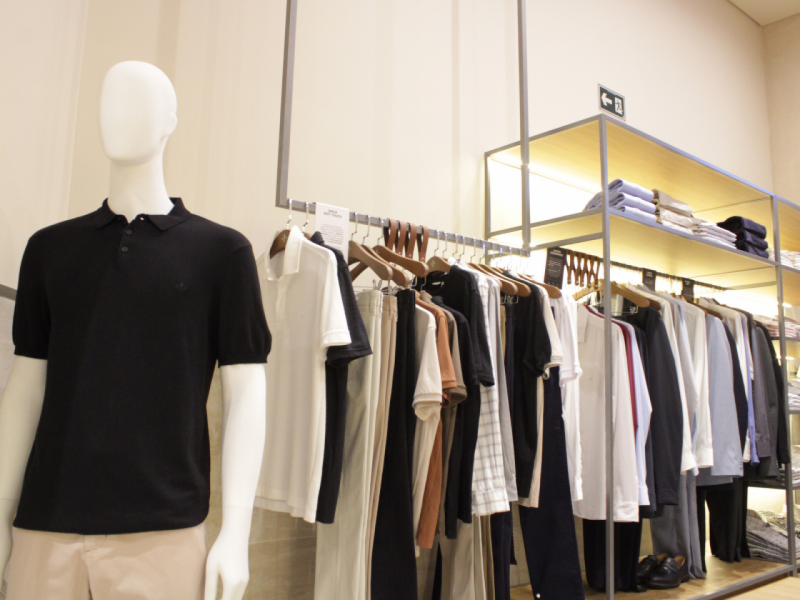 Manequim masculino de polo preta e prateleiras ao fundo com cabides de roupas escuras e camisas sociais dobradas no topo. 