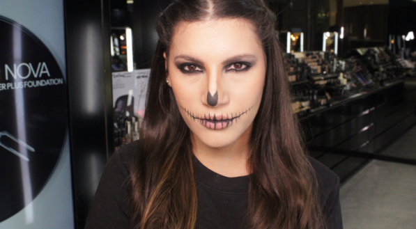 Um close-up frontal mostra a modelo com a maquiagem de caveira finalizada: esfumado preto, nariz e boca costurada. O fundo &eacute; o interior da loja MAC.