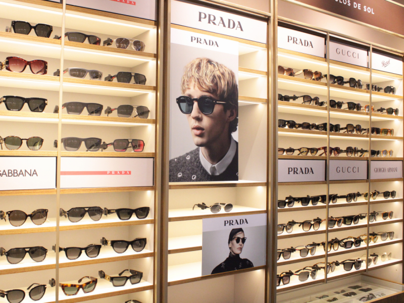 Exposição de óculos de sol em uma loja, com diversas prateleiras e caixas de marcas como Prada, Gucci e Persol, e pôsteres promocionais.