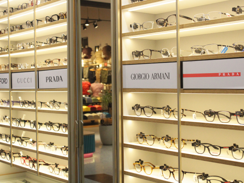 Prateleiras iluminadas de uma loja de óculos, exibindo diversos modelos de grau, com caixas de marcas como Prada, Gucci e Giorgio Armani.