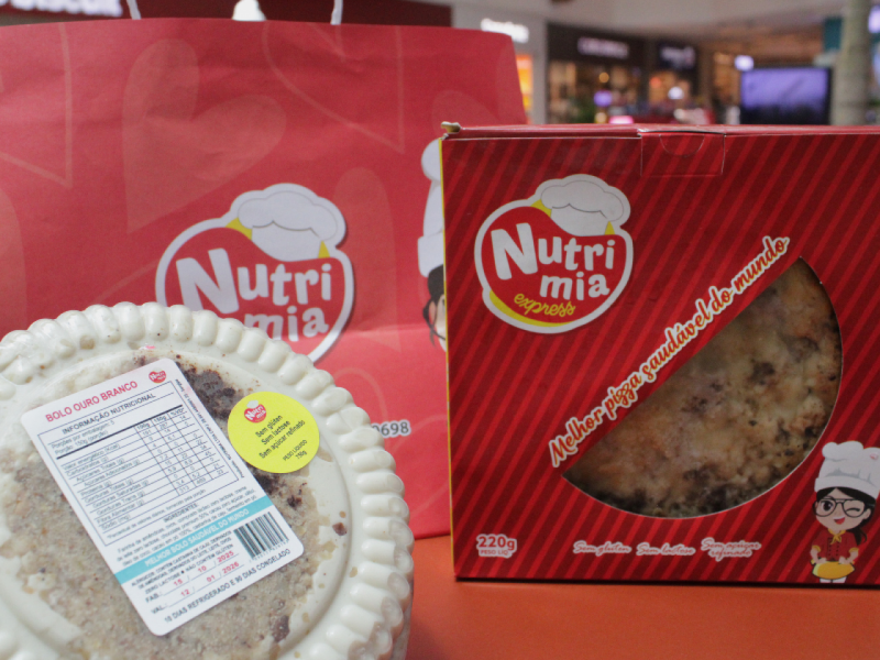 A imagem mostra um bolo saudável sabor ouro branco, uma pizza saudável e uma sacola da Nutrimia. A imagem mostra um bolo saudável sabor ouro branco, uma pizza saudável e uma sacola da Nutrimia.