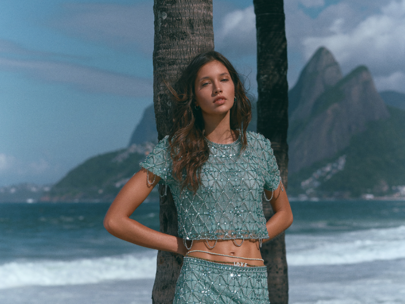Uma modelo com top cropped e minissaia turquesa cravejados de miçangas posa na praia, com coqueiros e o Morro Dois Irmãos ao fundo.