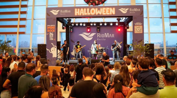 Uma banda de rock se apresenta em um palco em um shopping center durante um evento de Halloween, com uma grande plateia de adultos e crianças em frente. A decoração inclui o nome "RioMar Recife" e uma guirlanda festiva no alto.