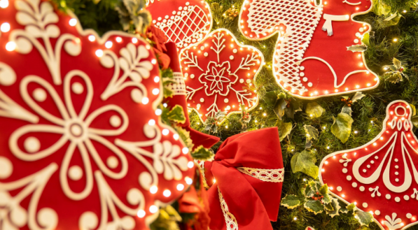 Uma foto em close de decorações natalinas em formato de biscoitos de gengibre vermelhos, iluminadas e detalhadas com padrões brancos de glacê. Um grande laço de fita vermelho com renda dourada é um elemento central.