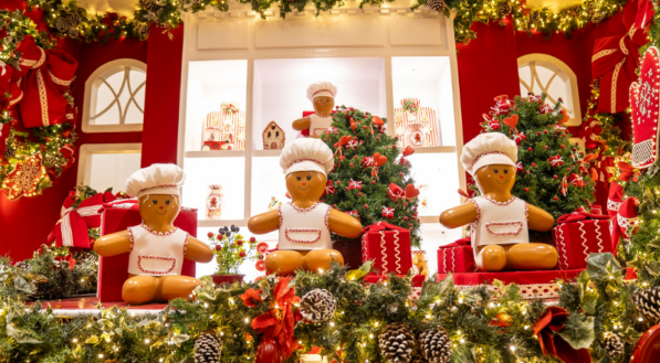 Um cenário natalino que simula uma cozinha com um forno grande no centro. Bonecos de homem-biscoito vestidos de chef estão sentados sobre presentes vermelhos e grandes talheres cenográficos, com mais bonecos visíveis dentro da tela do forno.