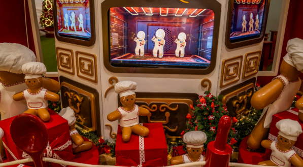 Um cenário natalino que simula uma cozinha com um forno grande no centro. Bonecos de homem-biscoito vestidos de chef estão sentados sobre presentes vermelhos e grandes talheres cenográficos, com mais bonecos visíveis dentro da tela do forno.