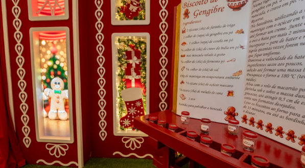 Uma parte da decoração vermelha e branca com janelas iluminadas e uma receita de Biscoito de Gengibre em destaque, escrita em português. Pequenos potes com ingredientes e um boneco de biscoito de gengibre em uma janela compõem a cena.