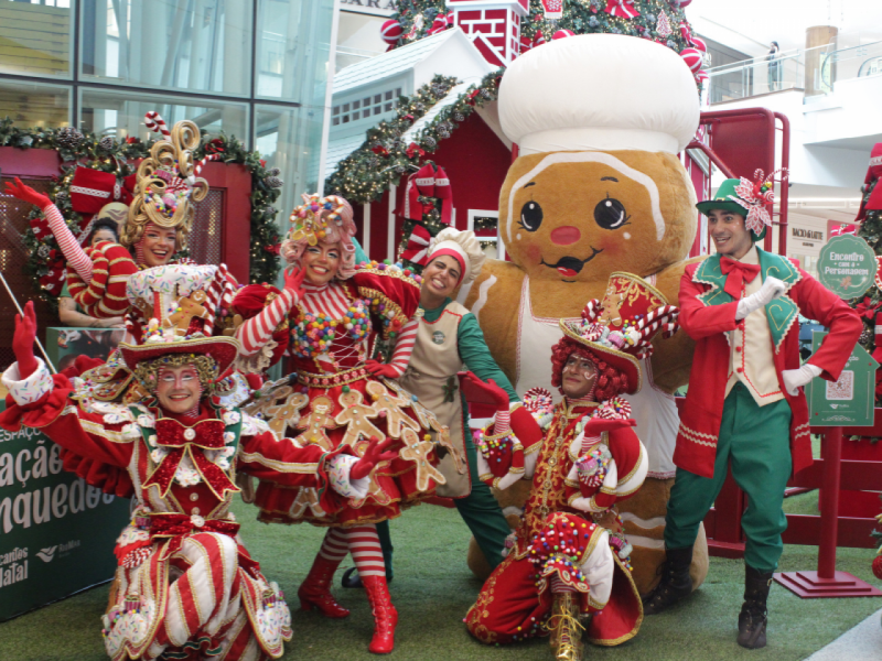 Um grupo de seis artistas fantasiados com trajes extravagantes e coloridos de tema natalino, inspirados em doces e biscoitos de gengibre, posa junto a uma grande mascote de Biscoito de Gengibre.