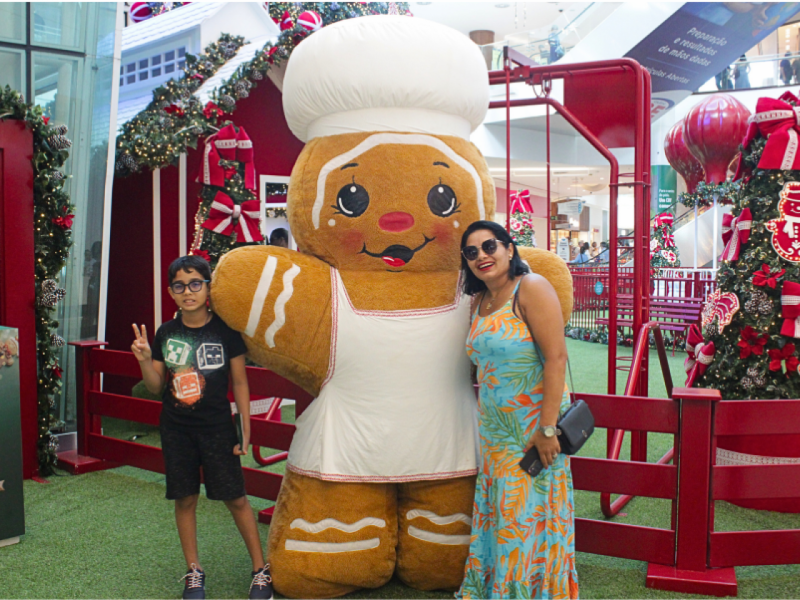Um menino e uma mulher posam com um grande mascote de Boneco de Gengibre vestido de chef em um cenário de Natal com casinha e cerca vermelhas.
