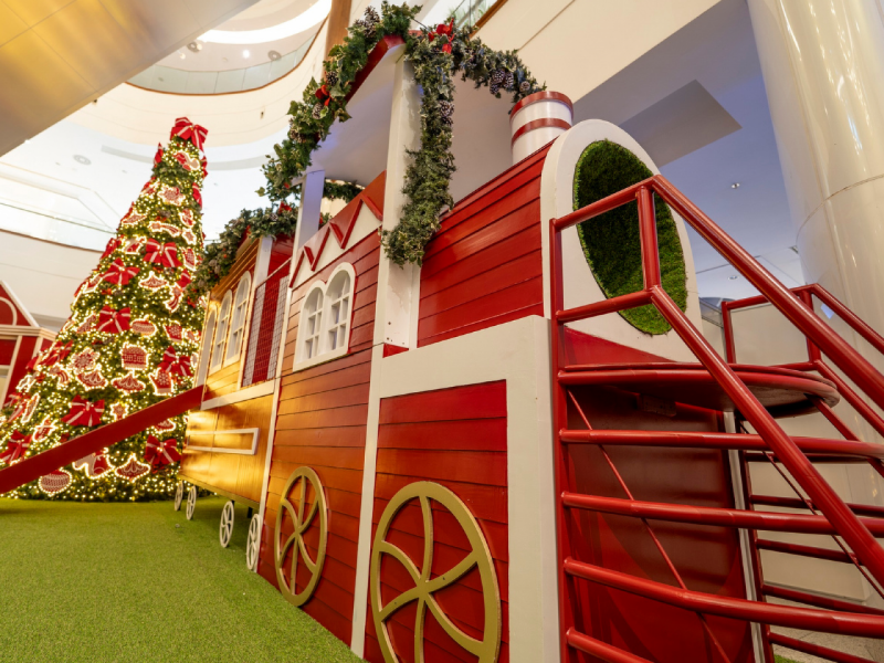 Uma locomotiva de trem cenográfica, grande e vermelha, que parece ser um escorrega ou área de brincar, decorada com guirlandas. Ao fundo, uma Árvore de Natal gigante e iluminada.
