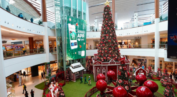 Uma vista panor&acirc;mica do &aacute;trio central de um shopping center, com uma enorme &aacute;rvore de Natal decorada em vermelho, verde e dourado, circundada por casinhas tem&aacute;ticas. A decora&ccedil;&atilde;o principal est&aacute; em uma &aacute;rea de piso verde que simula grama, ladeada por grandes esferas vermelhas, sob um elevador de vidro com um an&uacute;ncio digital.