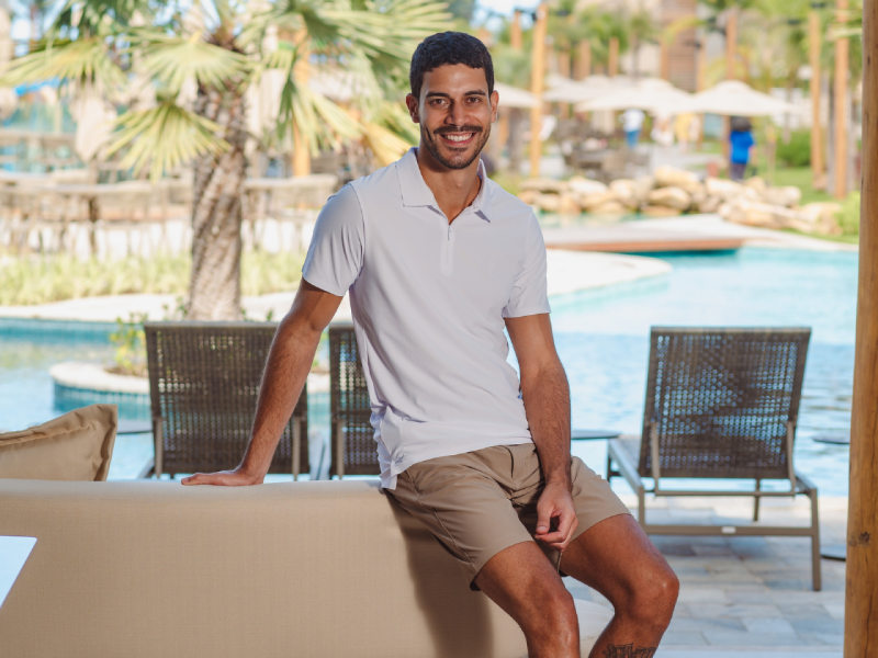Homem sorrindo, sentado à beira de um sofá perto de uma piscina e área de lazer. Veste uma polo branca de manga curta e shorts chino bege.