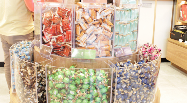 Exposi&ccedil;&atilde;o em forma de p&oacute;dio de guloseimas Kopenhagen, com divis&otilde;es cil&iacute;ndricas transparentes contendo chocolates coloridos. No fundo, um funcion&aacute;rio est&aacute; atr&aacute;s do balc&atilde;o de atendimento.