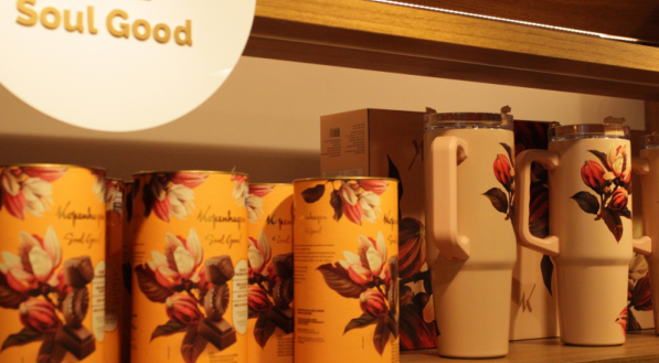 Close de uma prateleira de madeira clara exibindo latas cil&iacute;ndricas de chocolate com tema floral em laranja e canecas t&eacute;rmicas grandes e claras. Acima, um sinal de &quot;Soul Good&quot; &eacute; vis&iacute;vel.