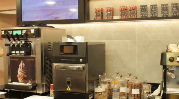 Vista da &aacute;rea de preparo de bebidas e sorvetes em uma loja Kopenhagen. O balc&atilde;o escuro exibe uma m&aacute;quina de sorvete soft, uma m&aacute;quina de caf&eacute; expresso e condimentos.