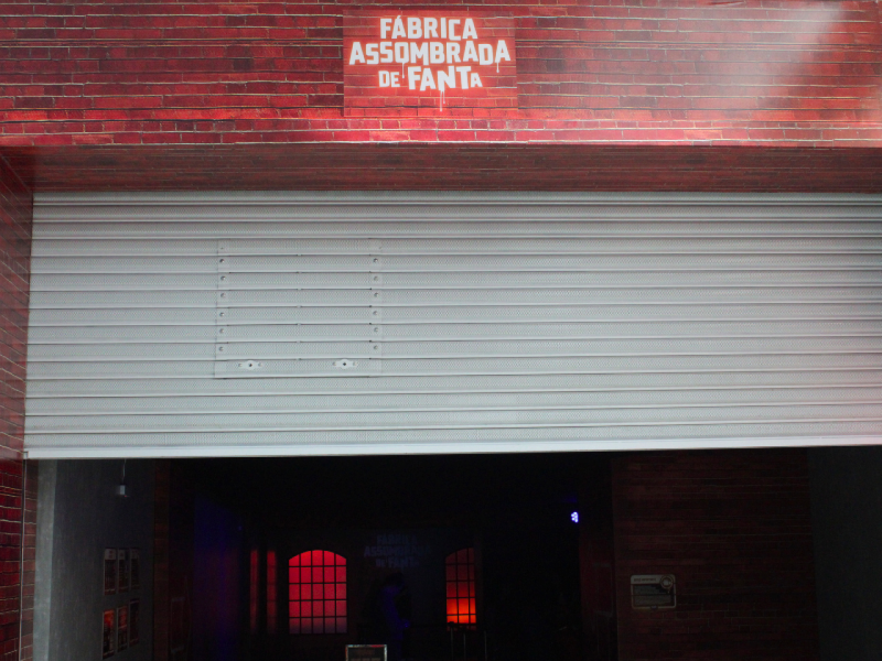 Uma grande porta de enrolar semiaberta em um prédio de tijolos vermelhos. O interior escuro revela janelas vermelhas iluminadas, reforçando o ambiente misterioso.