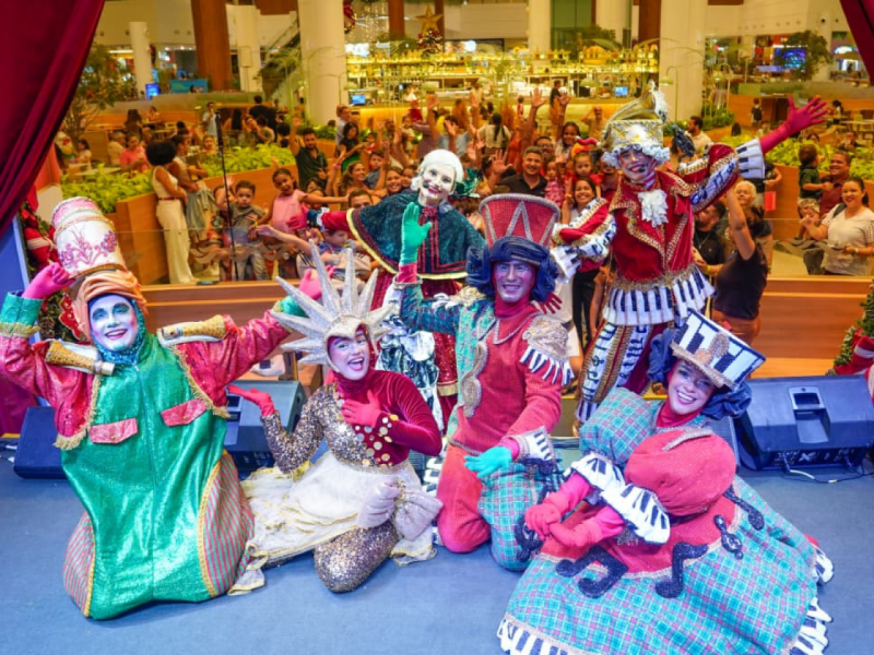 Um grupo de sete artistas fantasiados posa no palco de joelhos e agachados, sorrindo para a câmera, com a plateia e o cenário do shopping visíveis ao fundo.