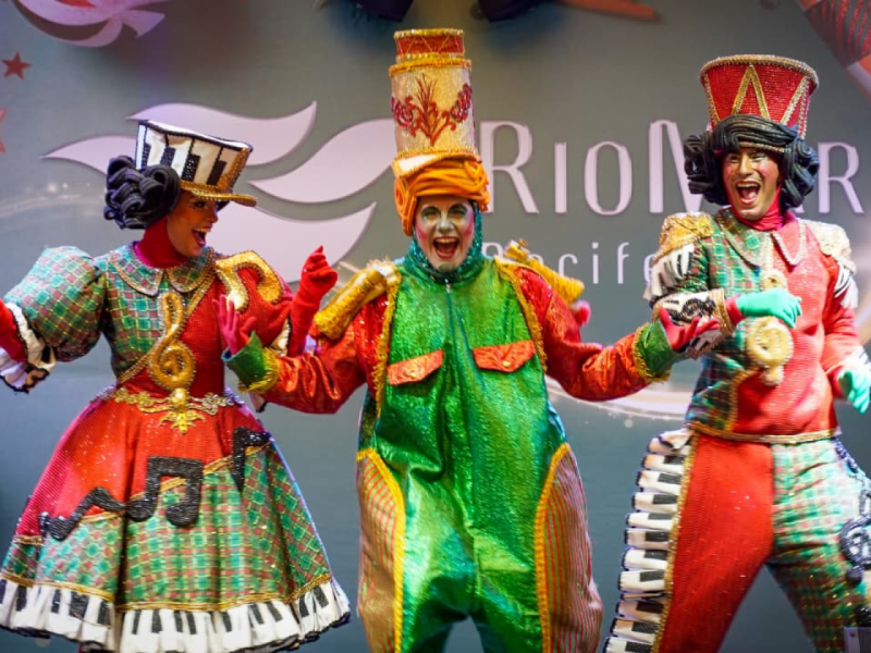 Três artistas em trajes vibrantes e temáticos, com destaque para a figura central estão em poses de alegria e performance no palco.