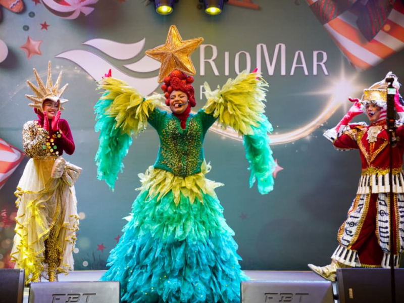 Três artistas em close-up, vestindo fantasias natalinas detalhadas de Árvore de Natal, notas musicais e estrela dourada, estão no palco em pose de performance.