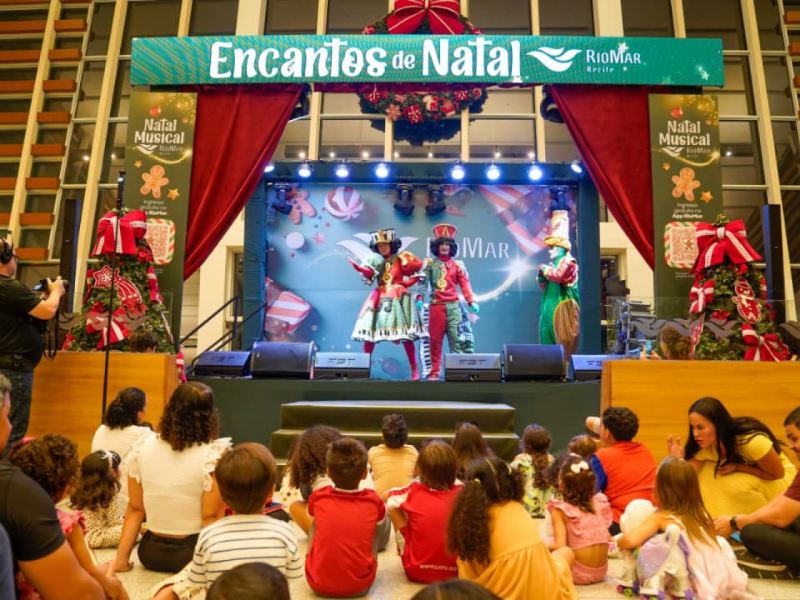 Três artistas fantasiados se apresentam no palco enquanto uma plateia, principalmente de crianças e seus pais, está sentada no chão da plateia, assistindo ao espetáculo.