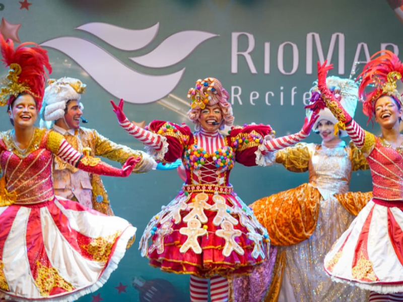 Artistas em trajes temáticos de doces e Natal se apresentando em um palco com o logo "RioMar Recife" ao fundo.
