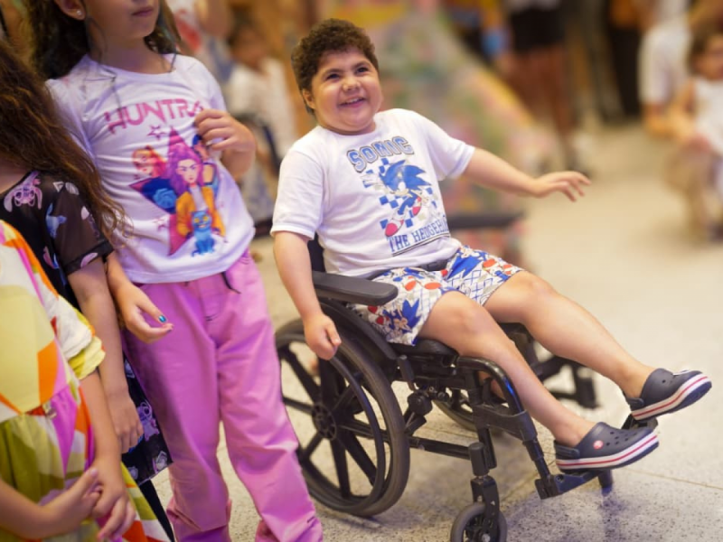 Um menino sorridente em uma cadeira de rodas, vestindo uma camiseta do Sonic, interagindo com outras crianças.
