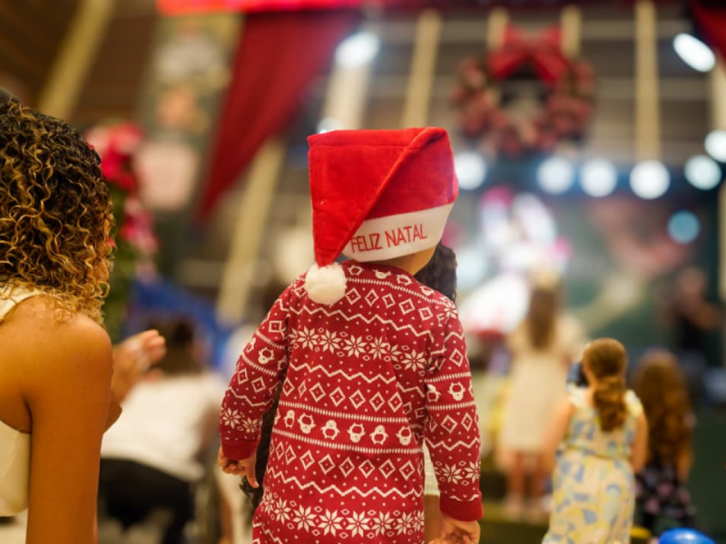 Uma criança, vista por trás, usando um gorro de Papai Noel com a inscrição "FELIZ NATAL" e um suéter vermelho de estampa natalina, assistindo a uma apresentação.