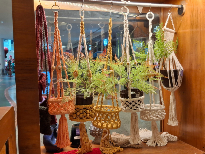 Uma prateleira de madeira escura exibindo vários suportes para plantas suspensos (macramê) em diferentes cores (laranja, amarelo, branco cru), com plantas verdes dentro de alguns deles.