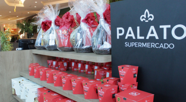 Vista de perto em um quiosque de supermercado, com um painel preto escrito &quot;PALATO SUPERMERCADO&quot;. Prateleiras de madeira exp&otilde;em v&aacute;rias caixas vermelhas de panetone e cestas de Natal embrulhadas em celofane com la&ccedil;os vermelhos no topo.