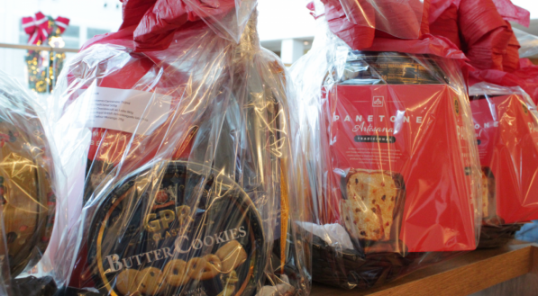 Foco em duas cestas de presentes de Natal na prateleira superior, embrulhadas em pl&aacute;stico transparente e fechadas com la&ccedil;os vermelhos. Uma mostra uma lata escura de biscoitos &quot;Butter Cookies&quot; e a outra uma caixa vermelha de panetone.