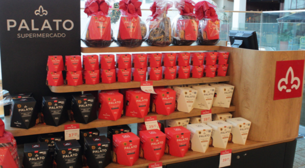 Vista frontal de um quiosque de madeira com o logo &quot;PALATO SUPERMERCADO&quot;. As prateleiras exibem simetricamente pilhas de caixas de panetone nas cores vermelha, preta e branca, e cestas de presentes no topo.
