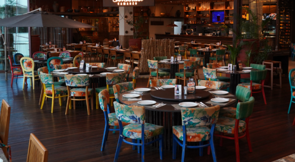 Interior do restaurante Camarada Camarão, com mesas e cadeiras de estampas florais e cores vibrantes, contrastando com o piso e o teto de madeira escura. Uma grande área externa com guarda-sol é visível no canto esquerdo.