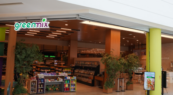 A fachada ampla da loja e empório "Greenmix" que vende produtos naturais e tem uma cafeteria. O interior exibe prateleiras recheadas de produtos, e no lado direito há um espaço de convivência com um painel de plantas.