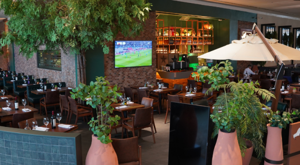 O restaurante Tapa de Cuadril, com uma área de mesas ampla e aconchegante, apresenta grandes vasos de plantas em terracota e árvores internas. O fundo é marcado por uma parede de tijolos e uma área de bar iluminada, com uma televisão ligada.