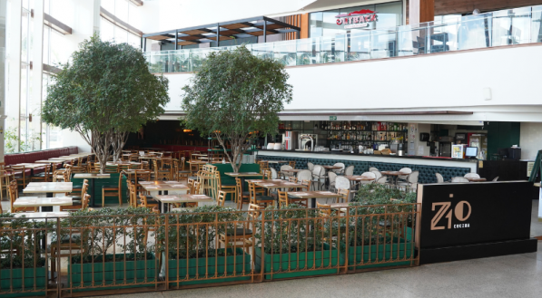 Vista frontal do restaurante Zio Cucina em um piso de shopping, com a área de mesas cercada por grades e arbustos verdes, e duas grandes árvores centrais. A entrada principal, à direita, tem o balcão do bar ao fundo.