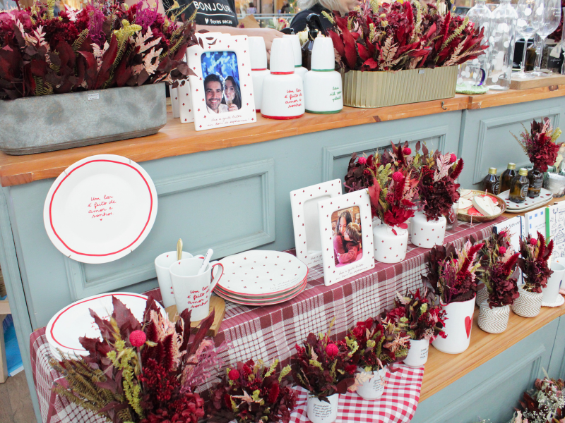 Visão geral de uma exposição de mesa com arranjos de flores secas vermelhas e marrons, louças brancas com detalhes vermelhos (estrelas, frases) e porta-retratos decorados.