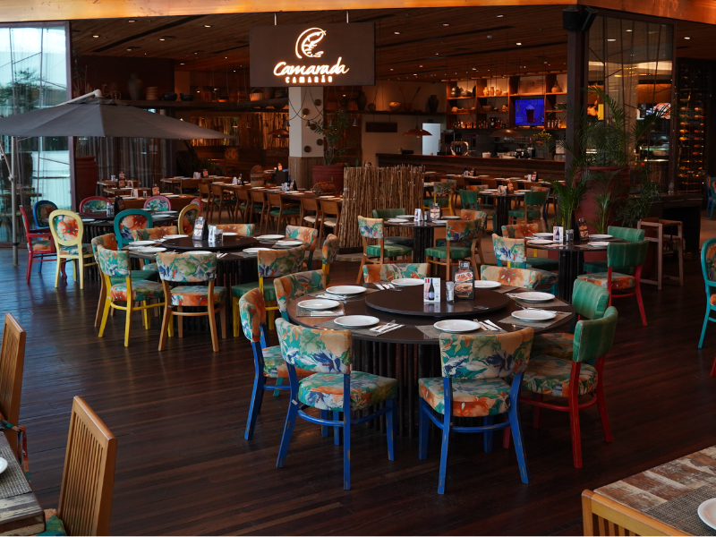 Interior do restaurante Camarada Camarão, com mesas e cadeiras de estampas florais e cores vibrantes, contrastando com o piso e o teto de madeira escura. Uma grande área externa com guarda-sol é visível no canto esquerdo.