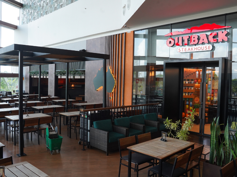O restaurante Outback Steakhouse tem sua área de mesas no corredor do shopping, com mesas de madeira rústica e escura, algumas sob uma pérgula preta. A entrada do restaurante, à direita, é marcada por um grande logotipo.