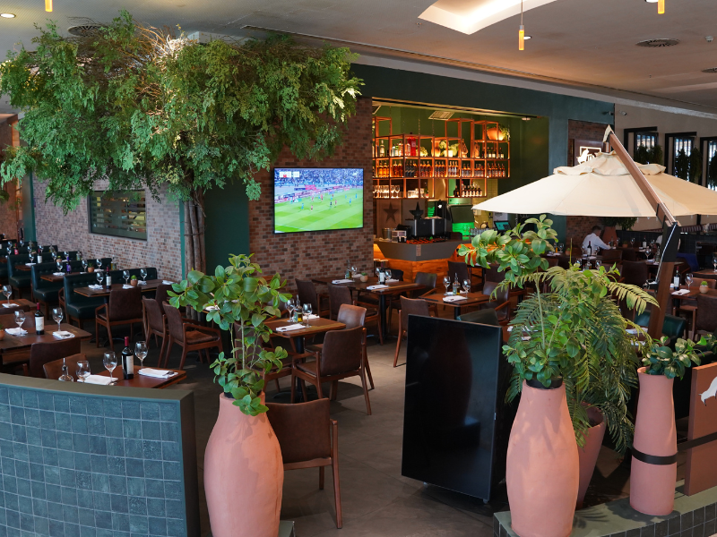 O restaurante Tapa de Cuadril, com uma área de mesas ampla e aconchegante, apresenta grandes vasos de plantas em terracota e árvores internas. O fundo é marcado por uma parede de tijolos e uma área de bar iluminada, com uma televisão ligada.
