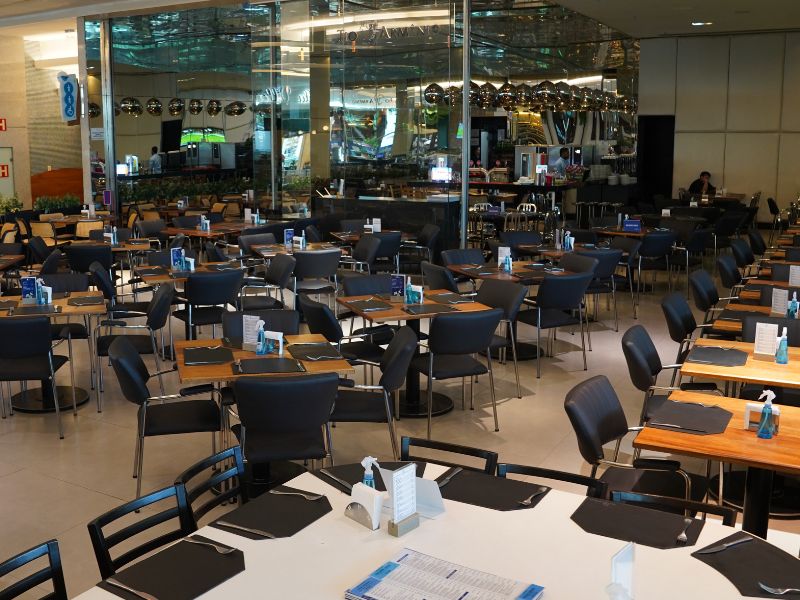 Visão interna do restaurante Tio Armênio, mostrando um salão com diversas mesas de tampo de madeira e cadeiras estofadas escuras, com itens de higiene sobre elas. O fundo do restaurante é marcado por uma grande parede de vidro espelhado.