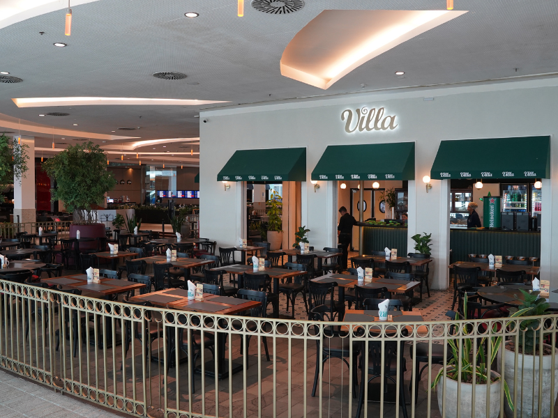 O Villa Bistrô, com sua fachada branca e toldos verdes esmeralda sobre as entradas, está cercado por uma grade de metal dourada. O salão tem mesas de madeira escura com cadeiras pretas e duas grandes árvores no interior.