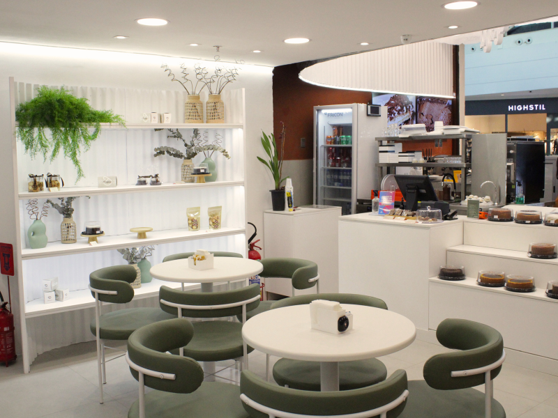 Vista da área de assentos moderna, com mesas redondas brancas e cadeiras de estofado verde-oliva. O ambiente é claro, com prateleiras decorativas e plantas à esquerda.